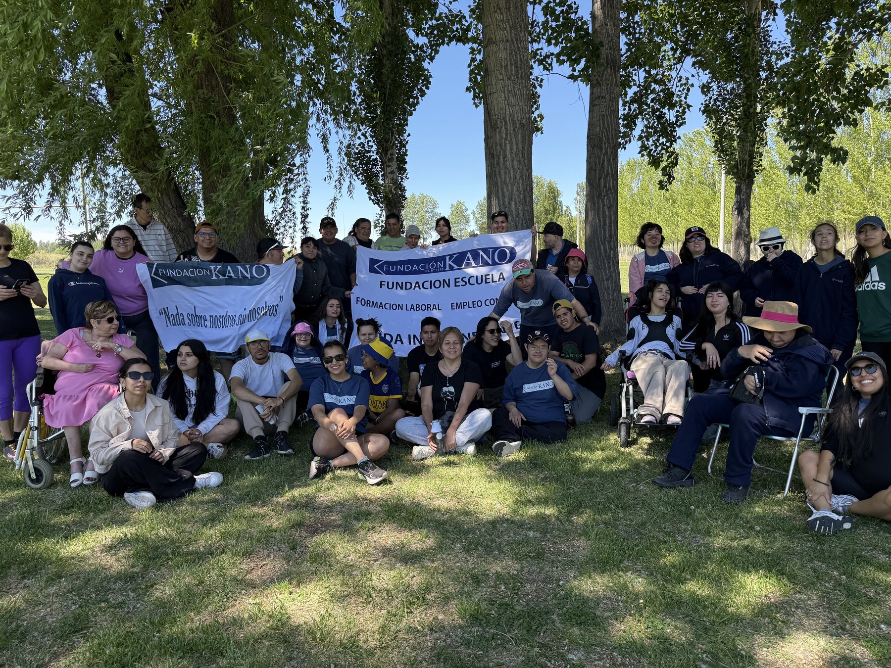 Alumnas, alumnos y equipo de Fundación Kano reunidos al aire libre bajo los árboles, sosteniendo una bandera de la fundación.